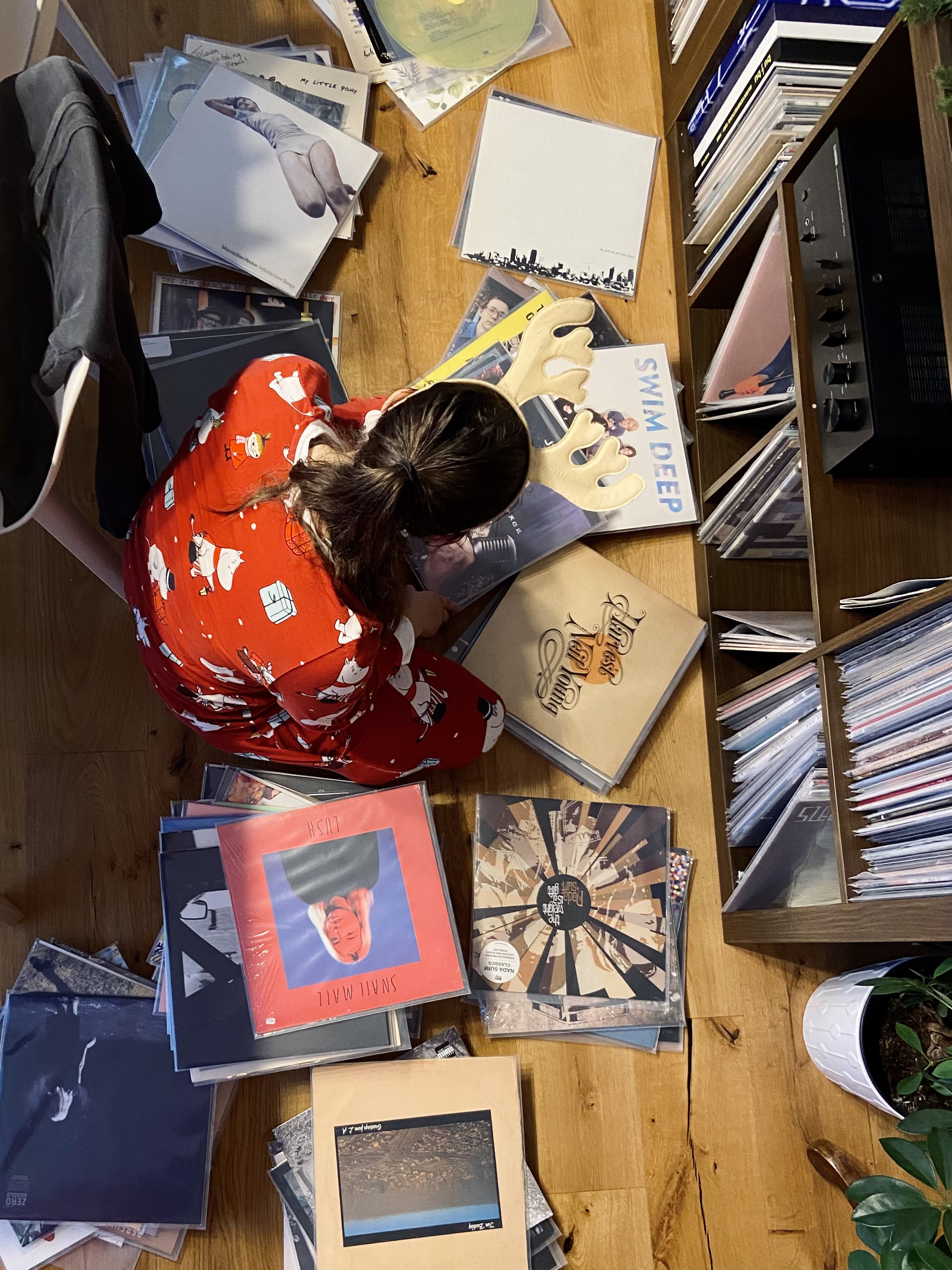 Woman wearing a Moomin Christmas pyjama and reindeer antlers, sitting on the floor surrounded by records like Neil Young's Harvest, Nada Surf's The Weight is a Gift, and Snail Mail's Lush