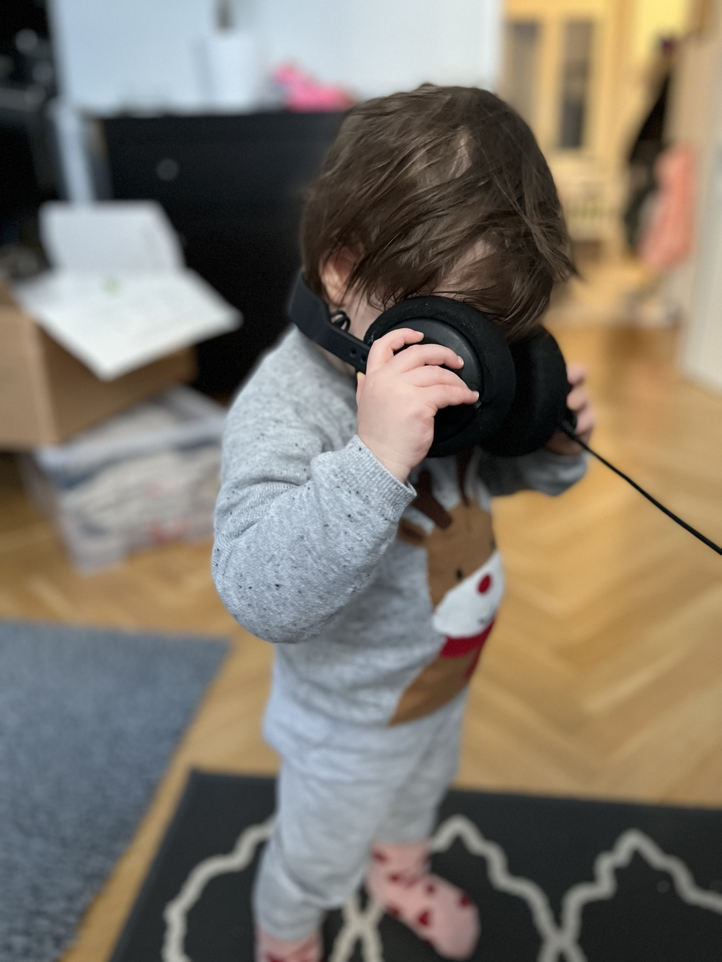 toddler wearing a reindeer sweater and standing, holding adult-sized headphones in front of her face