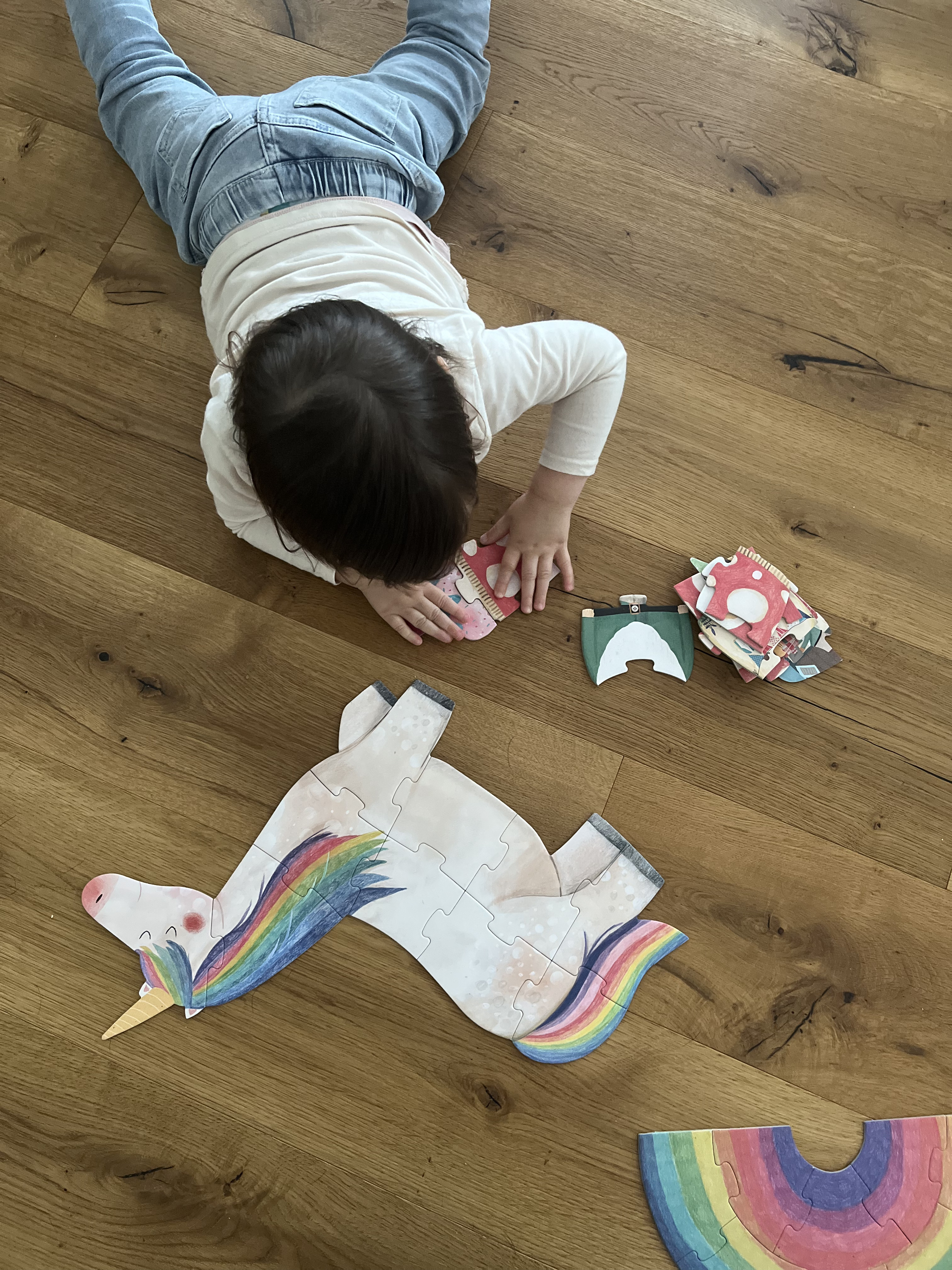 toddler laying belly-down on a hardwood floor and building a puzzle, with a completed unicorn-shaped puzzle in front of her