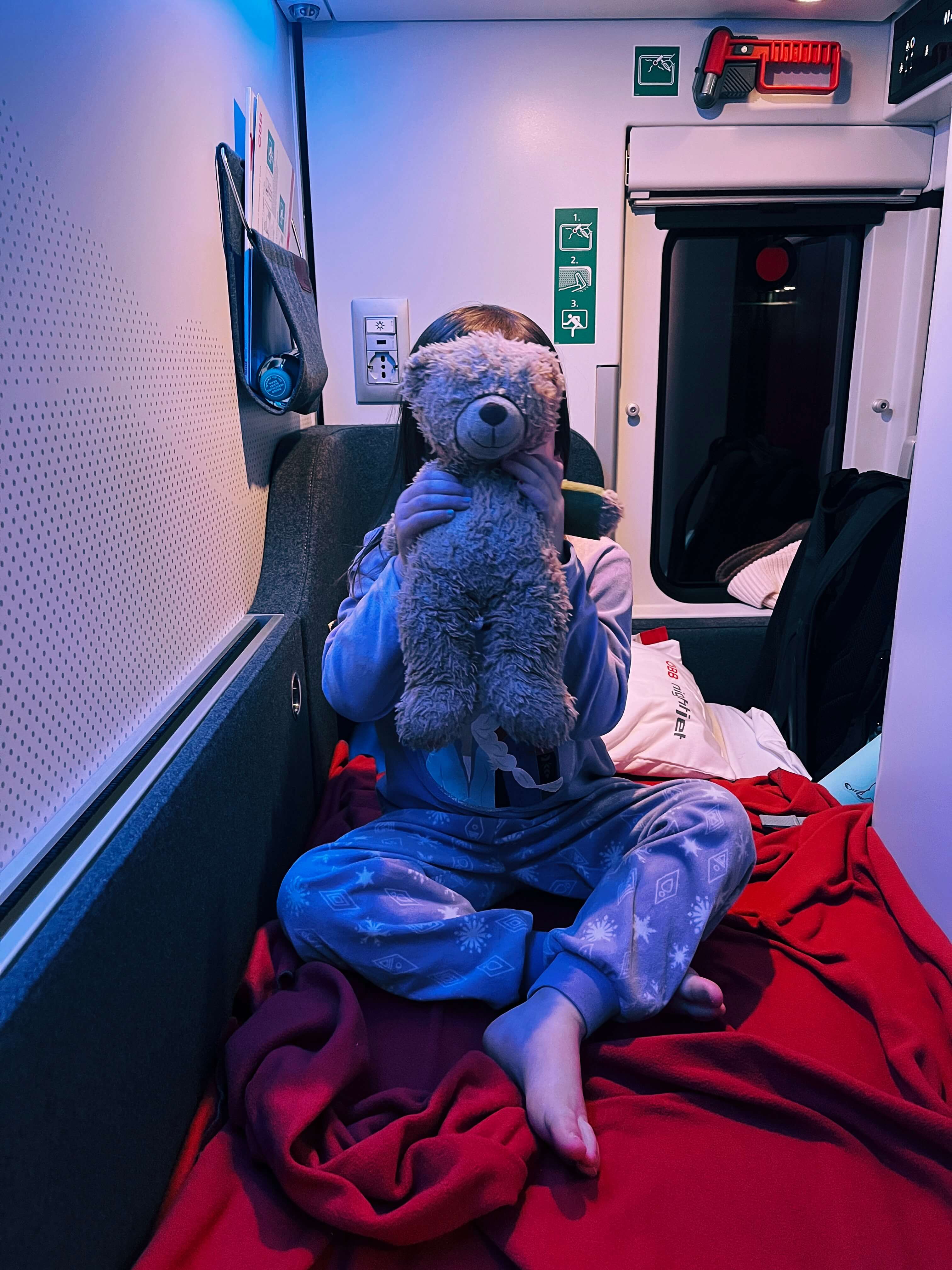 Child wearing purple pyjamas in a small train sleeper cabin, holding up a teddy bear in front of her face