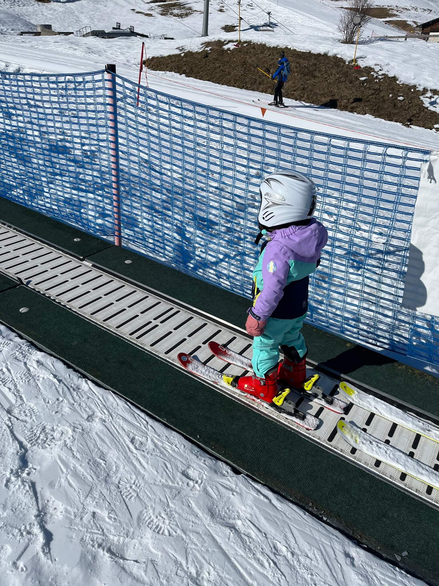 Child dressed in winter attire and wearing ski boots, a helmet and skis, standing on a moving walkway in a ski resort