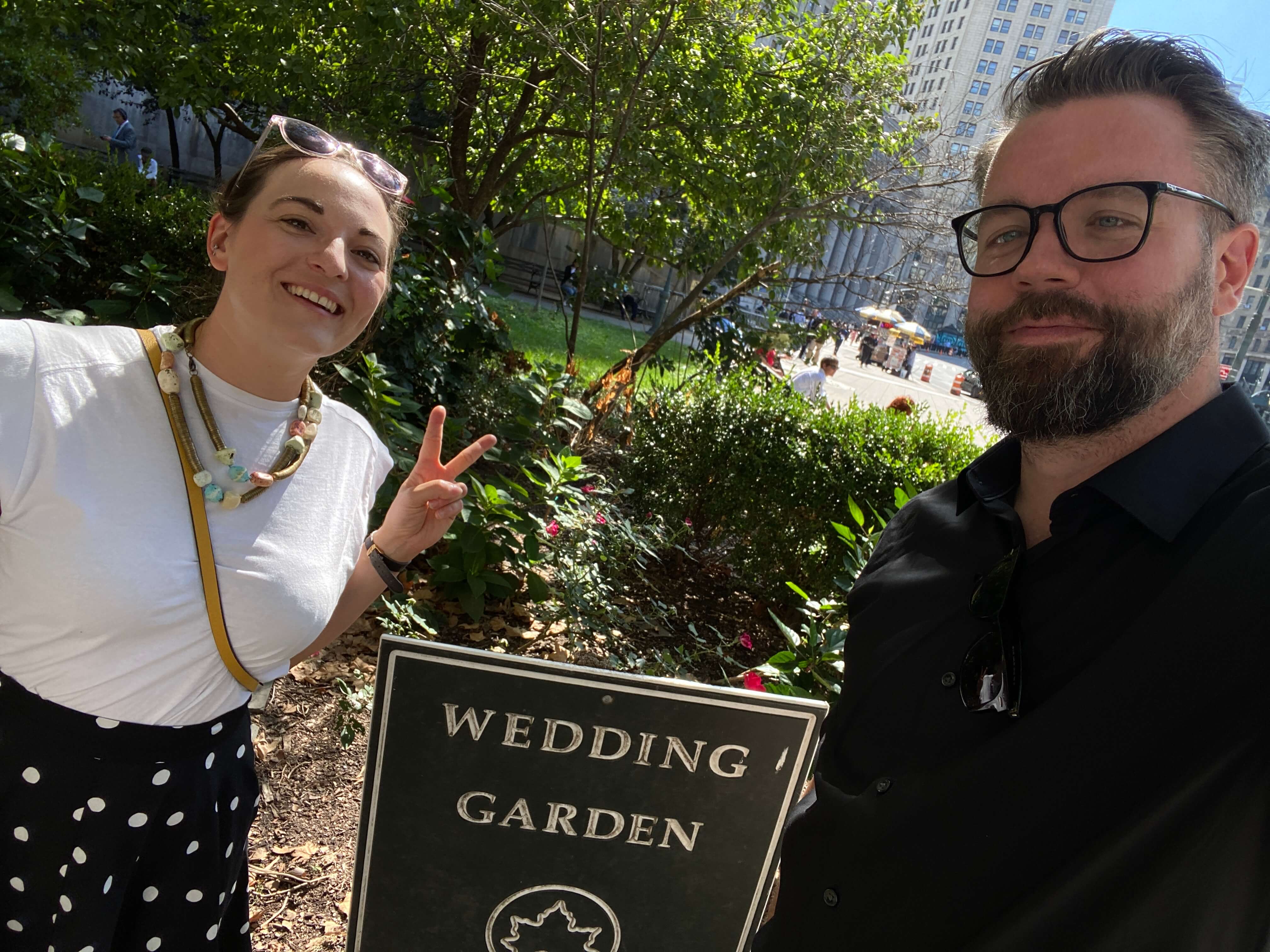 Woman and man, smiling at the camera, in front of a sign in a NYC park that reads 'Wedding Garden'